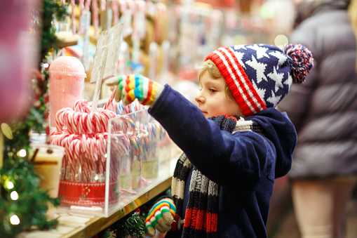 Little cute kid boy buying sweets from a cancy stand on Christmas market. Happy child on traditional family market in Germany. Preschooler in colorful winter clothes.