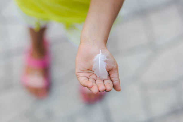 Beautiful close-up photography of a white bird feather in a little girl's hand. The open palm is in focus and the background is blurred, defocused, bokeh. The white feather is small. The hand is stretched, holding out the feather. The child is wearing green shirt and shorts, with pink sandals.  The photo is peaceful, tranquil. It awakes emotions of tenderness, love, innocence, memories of childhood, etc. The floor is grey stone. There is a lot of empty space, copy space, ideal for design, memes, backgrounds, animal posters, animal rights designs, etc.