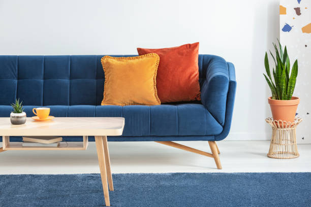 istock Orange and red cushions on a fancy, navy blue sofa and a basic, wooden coffee table on a blue rug in a white living room interior. Real photo. 1041057724