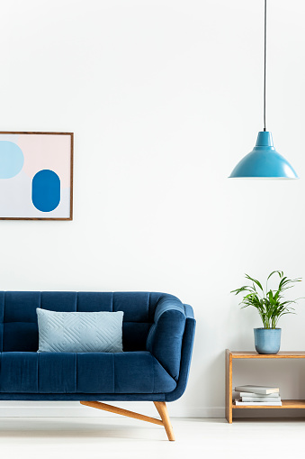 Retro bowl pendant light and a baby blue cushion on a dark, elegant sofa in a simple living room interior with white walls. Real photo.
