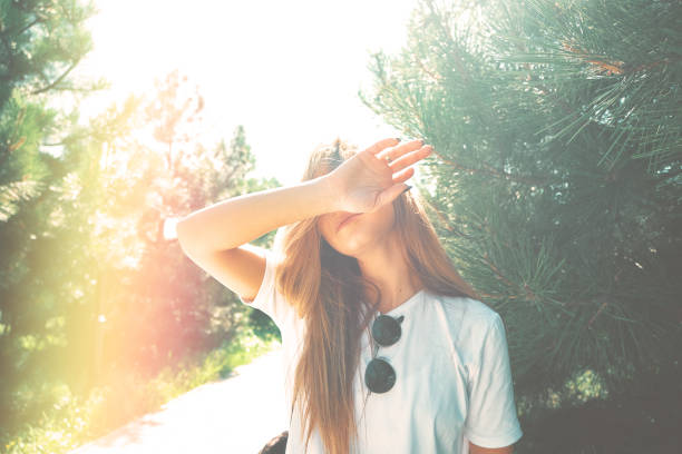 Close-up young woman with beautiful face features hide her face from sun rays with arm, pine in background. Light filters used.