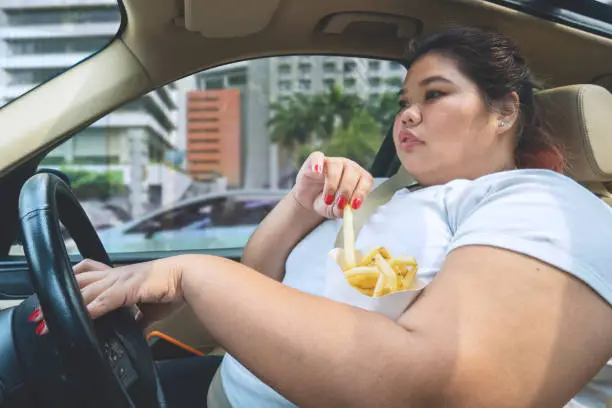 Image of overweight woman eating french fries during driving a car on the road