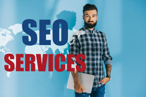 istock bearded developer in checkered shirt holding laptop, on blue with world map and SEO services 1041091502