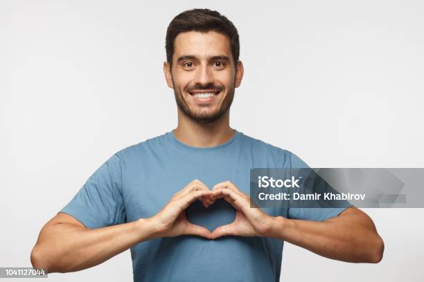 Retrato De Joven Sonriente Hombre En Azul Mostrando Camiseta Corazón Signo Aislado Sobre Fondo Gris Foto de stock y más banco de imágenes de Símbolo en forma de corazón