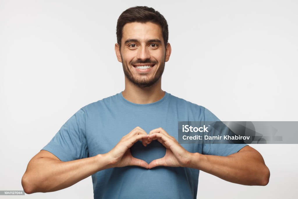 Retrato de jovem sorridente homem azul mostrando t-shirt coração signo isolado em fundo cinza - Foto de stock de Símbolo do Coração royalty-free