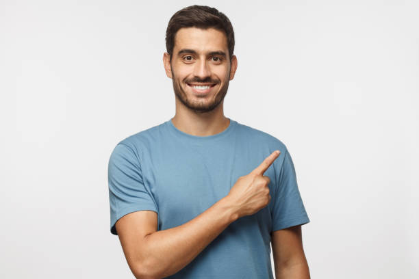 hombre joven en camiseta azul apuntando con su dedo aislado sobre fondo gris - aiming fotografías e imágenes de stock