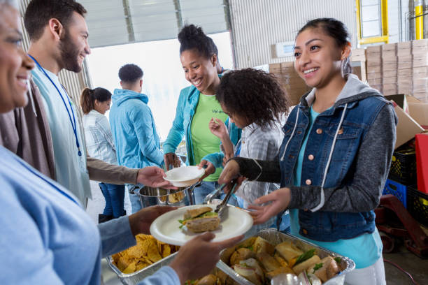 Group of volunteers serve meal at soup kitchen A group of cheerful volunteers, including a little girl, stand in a serving line at a local food kitchen and pass out soup, chips and sandwiches. community center food stock pictures, royalty-free photos & images