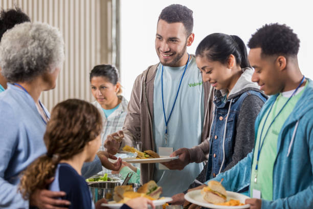 Young man enjoys serving meals at soup kitchen A young man stands in a volunteer serving line at his local soup kitchen and smiles as he passes out sandwiches. community center food stock pictures, royalty-free photos & images