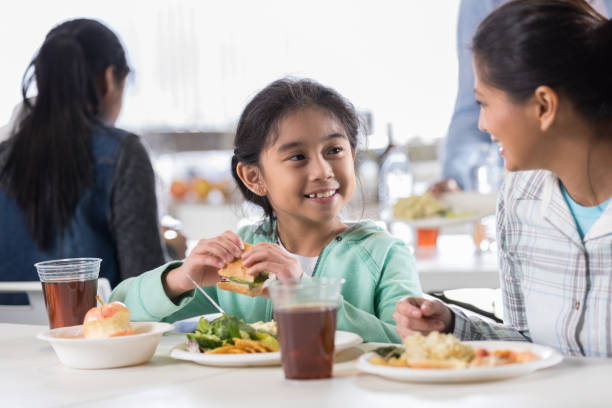 Little girl enjoys soup kitchen meal with mother A smiling elementary age little girl sits at a table with her mother and smiles at her as they enjoy a meal at their local soup kitchen. community center food stock pictures, royalty-free photos & images