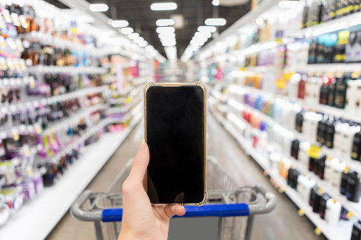 A human hand holds up a smart phone with a blank screen while shopping in a supermarket.  The shopper also pushes a cart down the shampoo aisle.