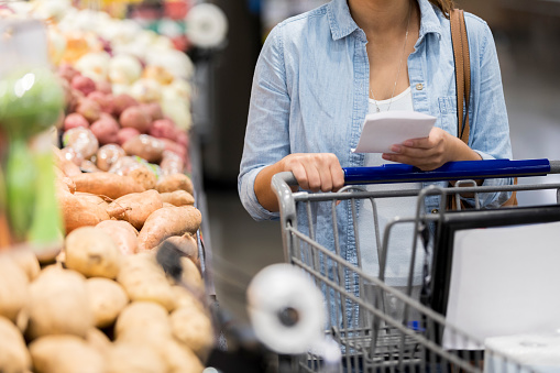 An unrecognizable woman pushes her cart past the potatoes in the grocery store.  She holds a shopping list.