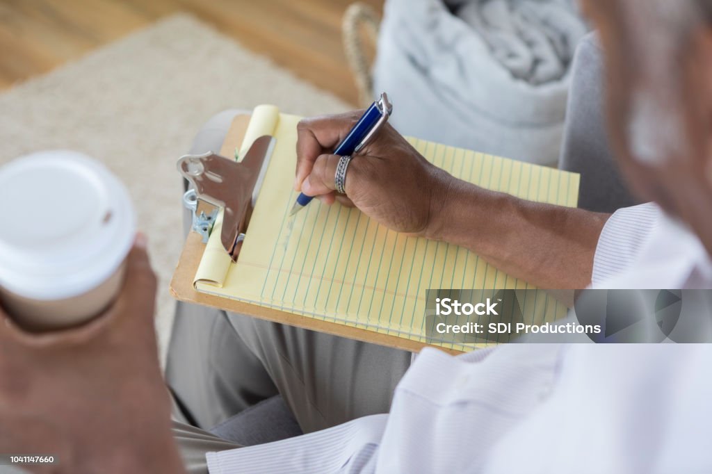 Unrecognizable man writes on yellow note pad High angle view of unrecognizable African American man writing on a yellow note pad. He is also holding a coffee cup. List Stock Photo