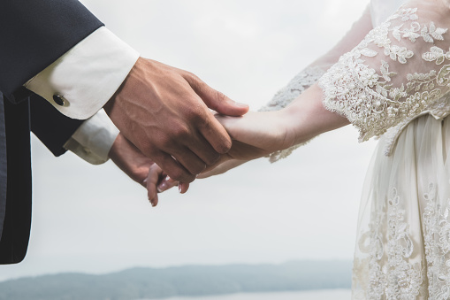 In this closeup, an unrecognizable bride and groom hold hands outdoors.  There is a scenic view in the background.