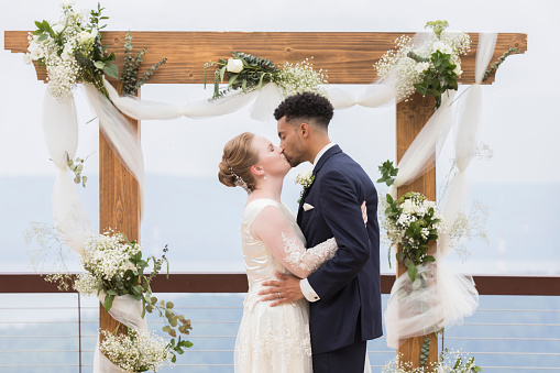 A bride and groom embrace and kiss in front of a decorated simple outdoor wedding altar.  They stand on a balcony in front of a scenic view.