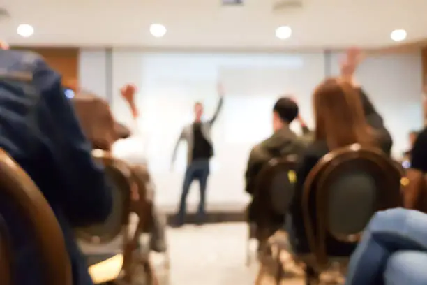 Photo of blurred group of people meeting in motivation seminar event at convention hall, speaker raising hand up and audience action follow , cheerful concept