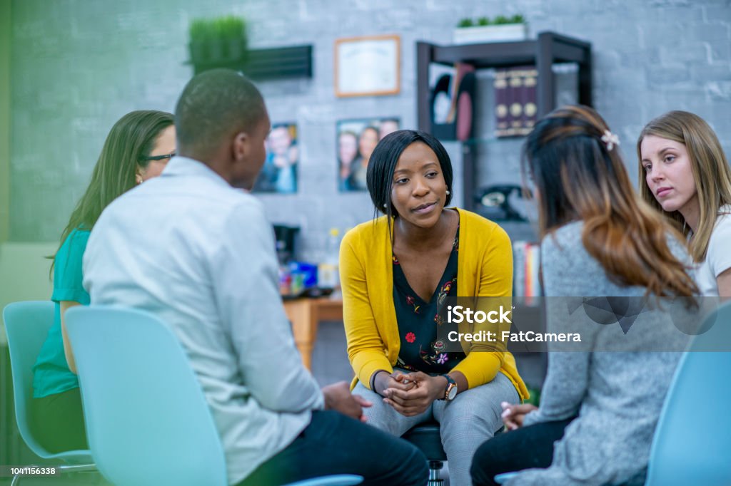 Group therapy session A diverse group of adults sit in a circle in their chairs and listen to and sympathize with each other with serious expressions. Group Of People Stock Photo