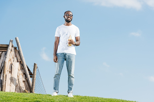 handsome african american man standing with soda in glass bottle on green hill