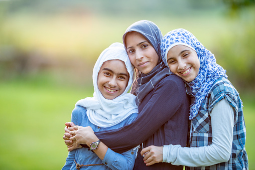 Three Muslim girls of different ages are outdoors in a park. They are embracing and smiling for the camera.