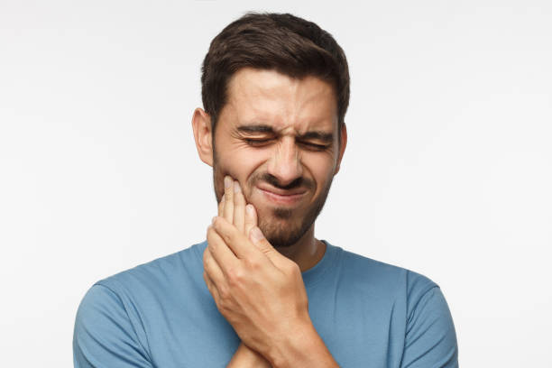 portrait de jeune beau mec solated sur fond gris, son visage déformé et les mains pressées pour joue comme il connaît mal de dent sévère et de soucis à ce sujet - rage de dents photos et images de collection