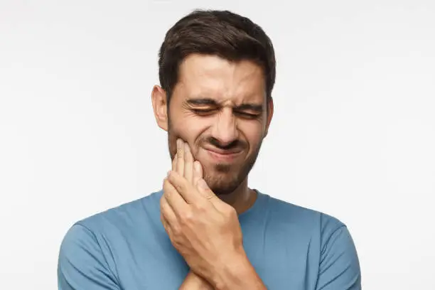 Portrait of young handsome guy solated on gray background, his face distorted and hands pressed to cheek as he is experiencing severe tooth ache and worries about it
