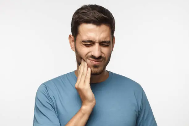 Tooth ache concept. Indoor shot of young male feeling pain, holding his cheek with hand, suffering from bad toothache, looking at camera with painful expression