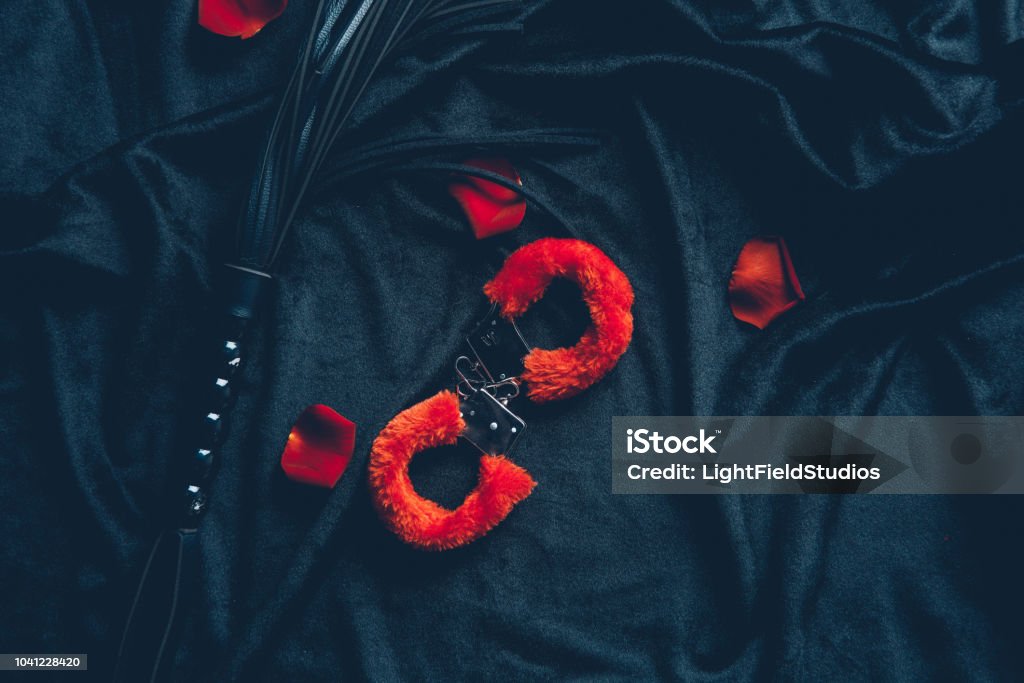 vue de dessus rouges moelleux menottes, fouet en cuir, des pétales de roses sur tissu noir - Photo de Menotte libre de droits