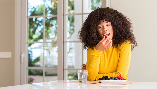 African american woman eating raspberries and blueberries at home with a confident expression on smart face thinking serious