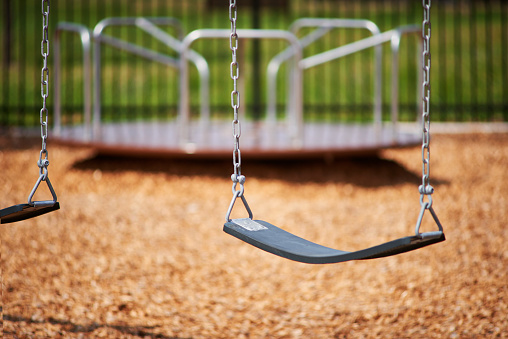 Image focused on a swing towards the right of the shot over a wood chip ground. There is a blurry merry-go-round in the back with a fence behind that. Green grass is shown blurry beyond the playground.