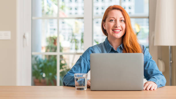 istock Redhead woman using computer laptop at home with a happy face standing and smiling with a confident smile showing teeth 1041333994
