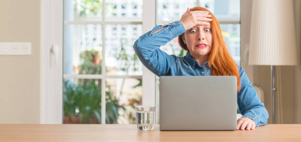 donna rossa che usa laptop per computer a casa stressata con mano sulla testa, scioccata dalla vergogna e dal viso a sorpresa, arrabbiata e frustrata. paura e turbamento per errore. - mistake foto e immagini stock