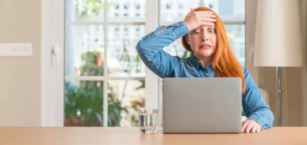 Redhead woman using computer laptop at home stressed with hand on head, shocked with shame and surprise face, angry and frustrated. Fear and upset for mistake.