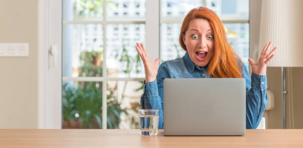istock Redhead woman using computer laptop at home very happy and excited, winner expression celebrating victory screaming with big smile and raised hands 1041334072