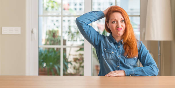 Redhead woman at home confuse and wonder about question. Uncertain with doubt, thinking with hand on head. Pensive concept.