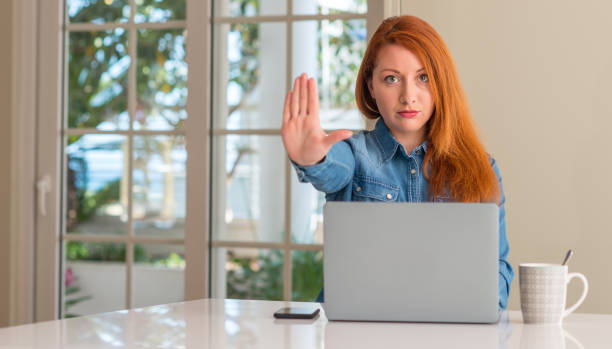 istock Redhead woman using computer laptop at home with open hand doing stop sign with serious and confident expression, defense gesture 1041349130