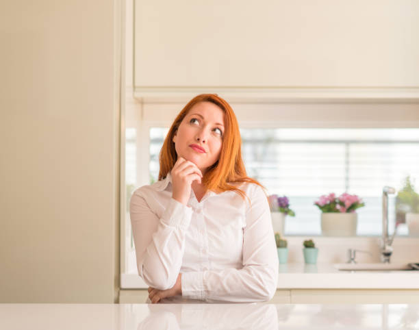 istock Redhead woman at kitchen with hand on chin thinking about question, pensive expression. Smiling with thoughtful face. Doubt concept. 1041351978