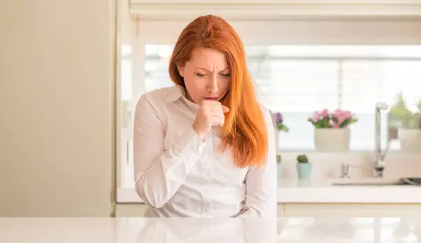Photo of Redhead woman at kitchen feeling unwell and coughing as symptom for cold or bronchitis. Healthcare concept.