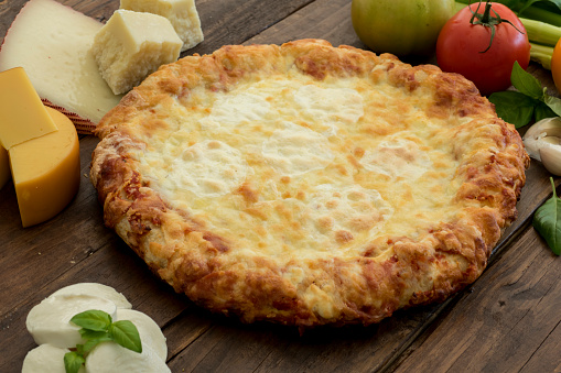 Cheese pizza on a rustic wooden table, surrounded by its ingredients: different kind of cheeses (mozzarella, provolone, parmigiano and cheddar), tomato, garlic and basil. Studio photography. High angle view. Horizontal composition with copy space. This image evokes the true tradition of homemade rustic pizza with natural and fresh ingredients.