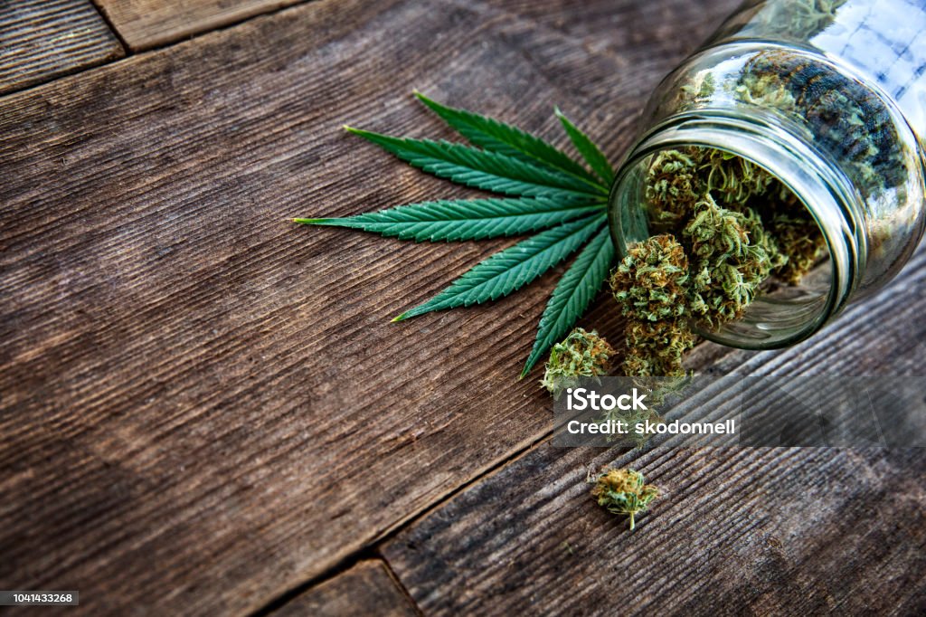 Cannabis bud pouring out of a glass jar on wood background This is a stock photograph involving cannabis, marijuana and its implications in America has just slowly been legalized and used for medicinal and medical purposes and what that means to our economy and culture. Cannabis - Narcotic Stock Photo