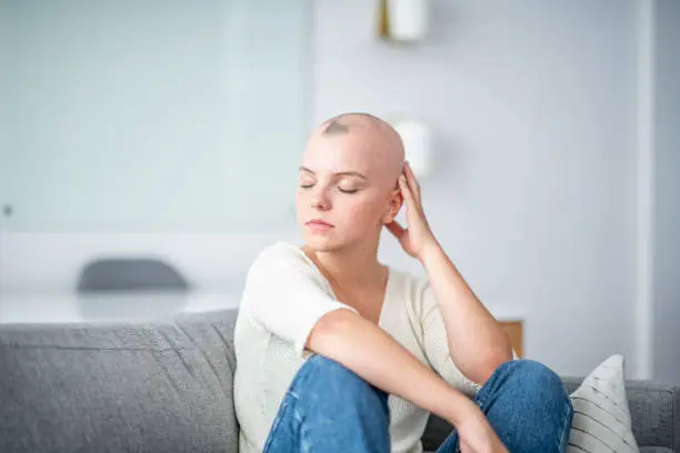 A woman is indoors in her living room. Her head is shaved due to chemotherapy. She is sitting on the couch with her eyes closed.