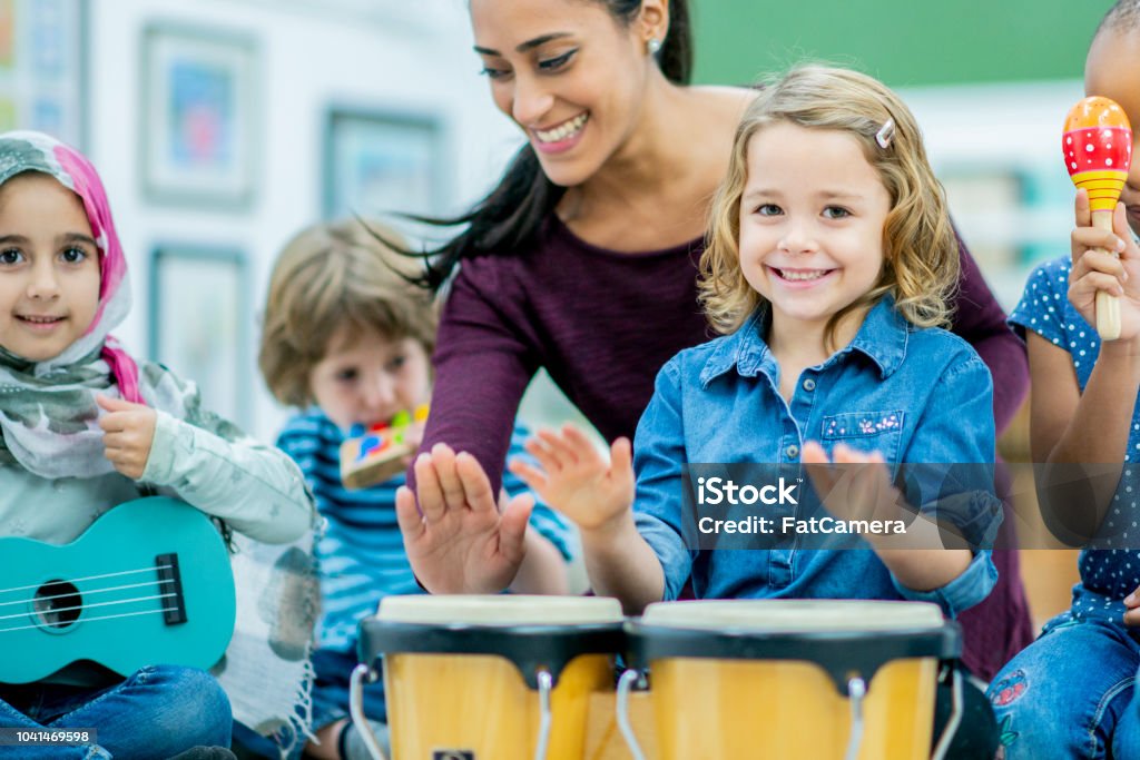 Having fun playing the hand drums in elementary school A diverse group of students plays musical instruments in preschool class with their teacher, who is smiling. Music Stock Photo