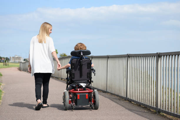 Boy in wheelchair holding mother's hand on path Rear view of mid adult mother holding hand of 6 year old son with muscular dystrophy, operating a wheelchair, and walking along coastal path essex england photos stock pictures, royalty-free photos & images