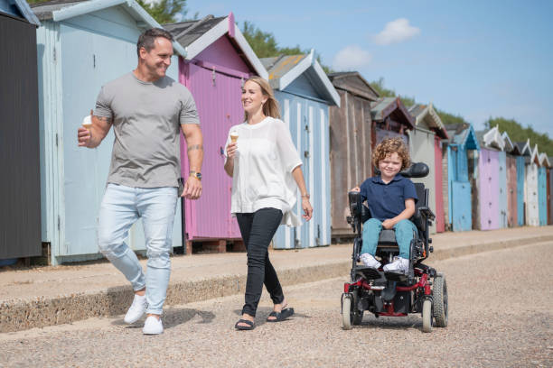 Young boy controlling wheelchair on day out with parents 6 year old boy with muscular dystrophy operating his wheelchair on day out to the seaside with mid adult mother and mature father clacton on sea stock pictures, royalty-free photos & images