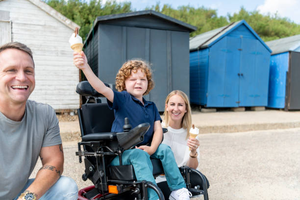 Boy in wheelchair holding up ice cream with parents, by beach huts Portrait of young boy with muscular dystrophy, sitting in wheelchair at the coast, enjoying ice cream treat with mid adult mother and mature father, looking towards camera and smiling clacton on sea stock pictures, royalty-free photos & images