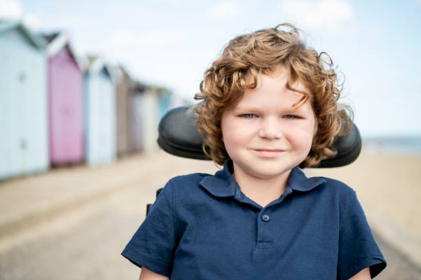 Portrait of 6 year old boy in wheelchair at British seaside Young boy with muscular dystrophy, sitting in his wheelchair on day out at the seaside, looking at camera, smiling and cheerful clacton on sea stock pictures, royalty-free photos & images