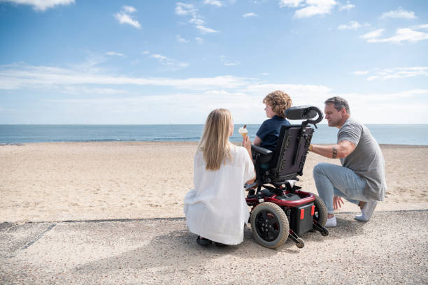 Parents crouching by son in wheelchair at beach Mid adult mother and mature father kneeling by 6 year old child with muscular dystrophy by the beach, enjoying an ice cream while looking at the view. Clacton on Sea. essex england photos stock pictures, royalty-free photos & images