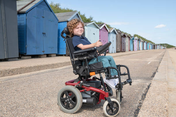 Boy with disability sitting in wheelchair by beach huts at seaside Full length, side view portrait of 6 year old disabled boy on summer vacation, looking at camera, smiling and cheerful clacton on sea stock pictures, royalty-free photos & images