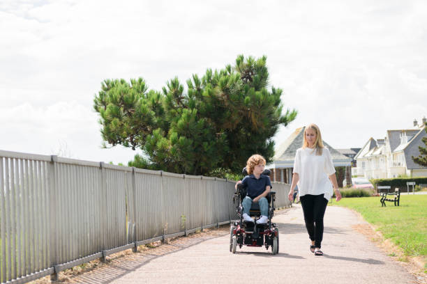 Mother walking beside son in wheelchair Front view of mid adult mother walking in the sunlight beside her 6 year old son with muscular dystrophy, operating his wheelchair clacton on sea stock pictures, royalty-free photos & images