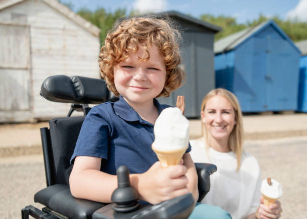 Disabled boy eating ice cream at beach with mother Mid adult mother enjoying ice cream treat by the seaside with her 6 year old son with muscular dystrophy in wheelchair, looking towards camera,  smiling and cheerful, close up essex england photos stock pictures, royalty-free photos & images