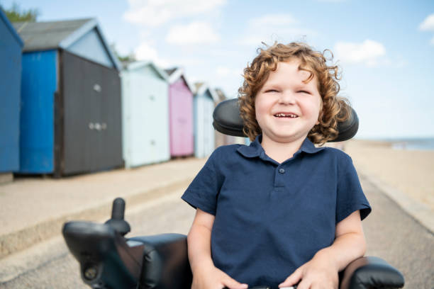 Portrait of 6 year old boy with disability laughing seaside Boy with muscular dystrophy sitting in wheelchair at the seaside, smiling and laughing with mouth open, looking happy and cheerful essex england photos stock pictures, royalty-free photos & images