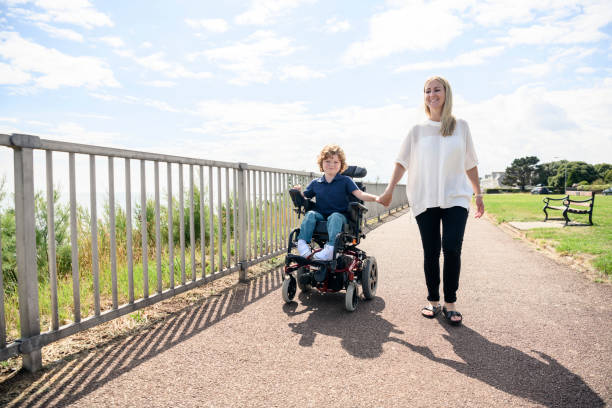 Disabled boy in wheelchair holding mother's hand on path Portrait of 6 year old disabled boy with muscular dystrophy, operating electric wheelchair while holding hands with his mid adult mother in the sunlight essex england photos stock pictures, royalty-free photos & images